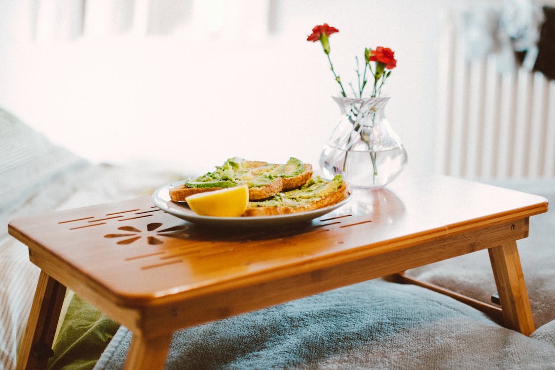 Free A cozy breakfast setting featuring avocado toast on a wooden tray with flowers in a vase. Stock Photo