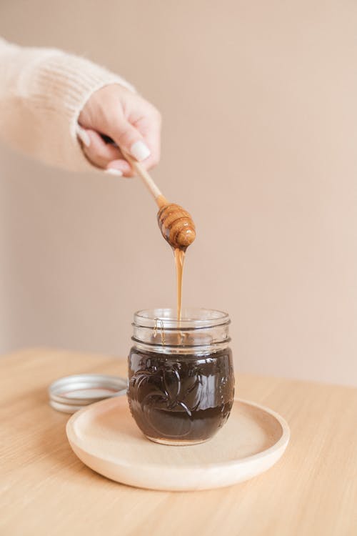 Free A hand holding a honey dipper drizzles honey into a jar on a wooden plate indoors. Stock Photo
